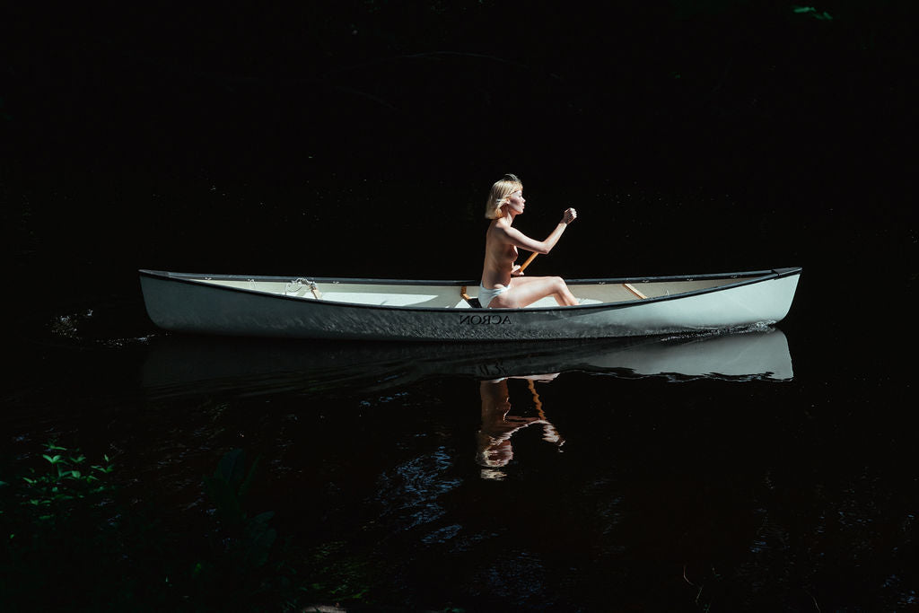 Nude girl paddles a blue canoe through a dark forest